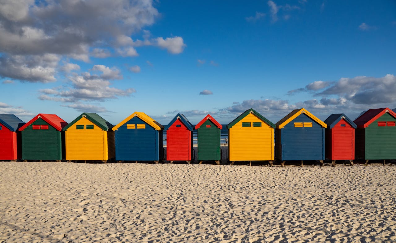 our-services-1 A row of vibrant beach huts on the sandy shores of Muizenberg, South Africa under a bright blue sky.